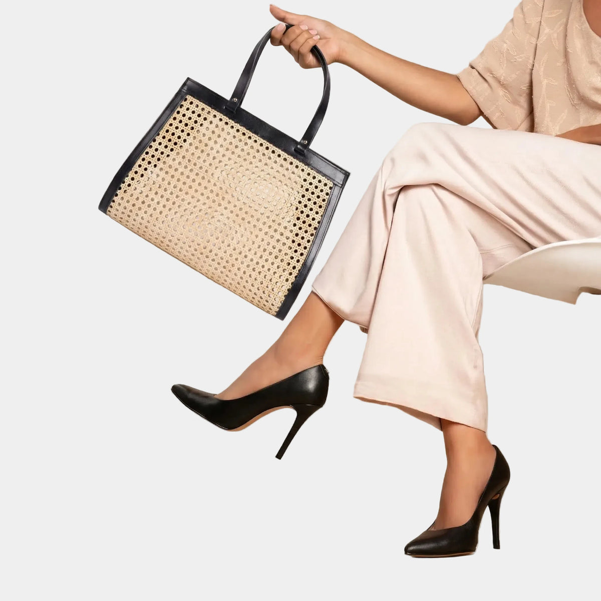 Model seated, holding the Lamia rattan cane tote with black leather handles