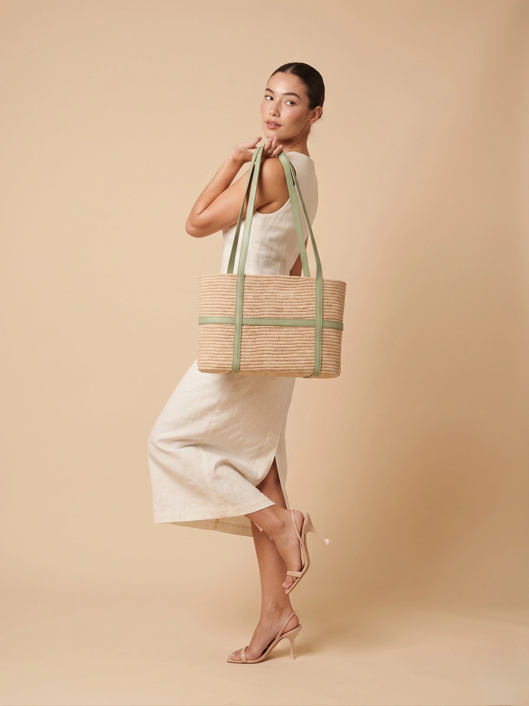 Woman in white dress carrying Kenza structured raffia tote with sage leather shoulder straps