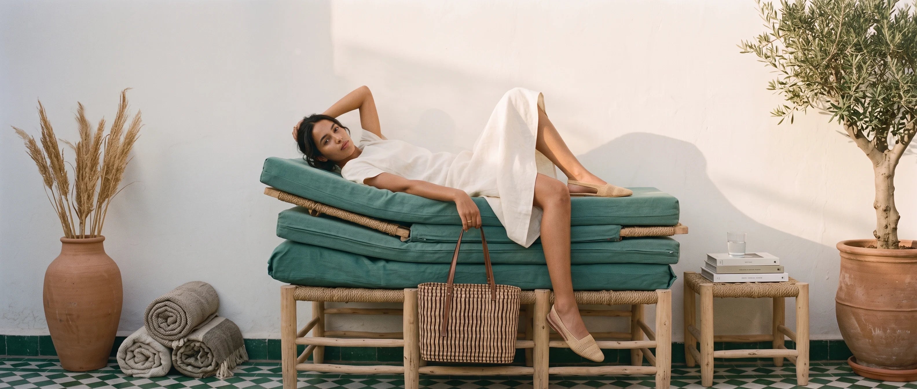 A woman in a white dress reclines on a teal cushioned daybed with bamboo legs, styled in a bright room with a wicker handbag, rolled towels, a terracotta vase with dried grass, and a potted olive tree.
