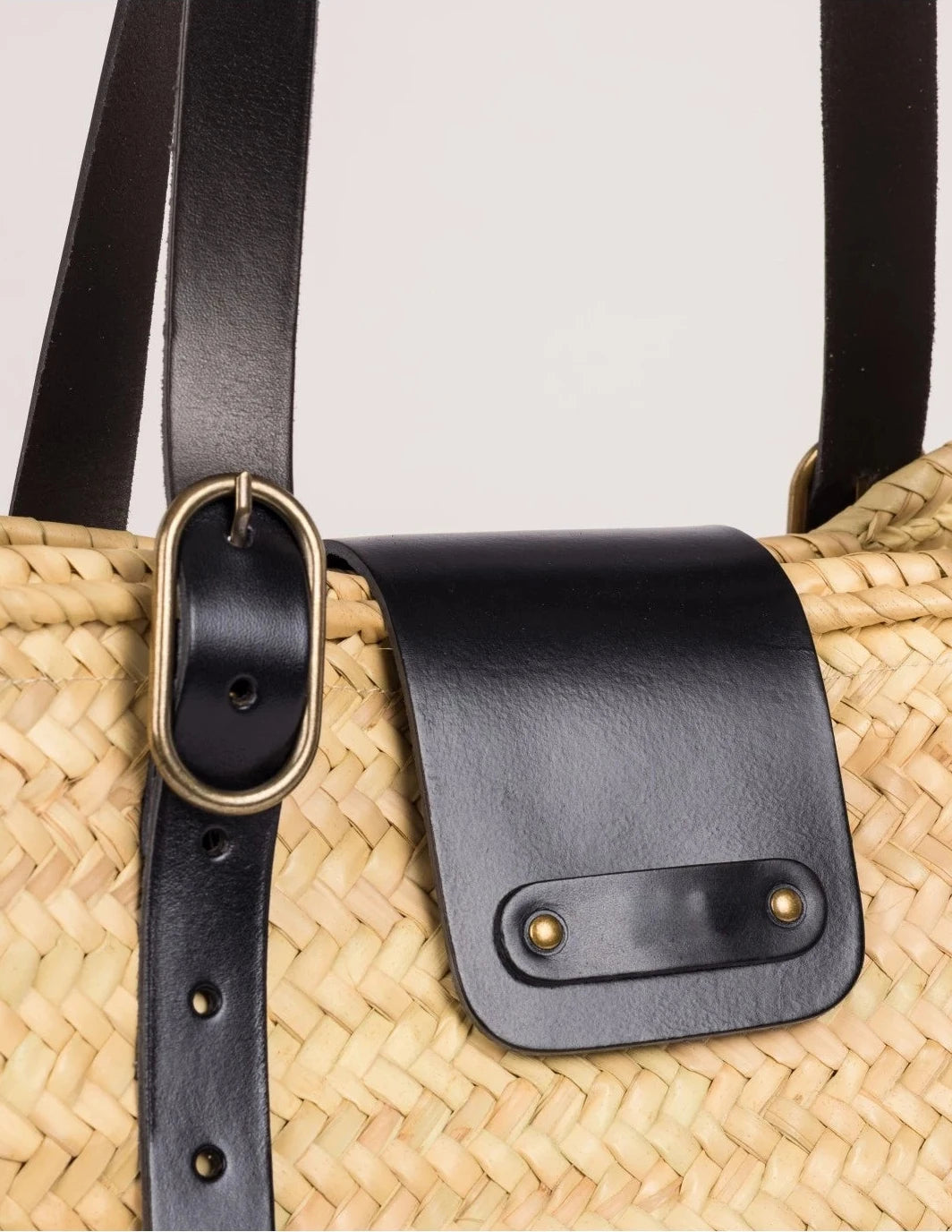 Close-up of Aicha basket bag showing black calfskin leather flap, buckle strap, and woven palm leaf texture.