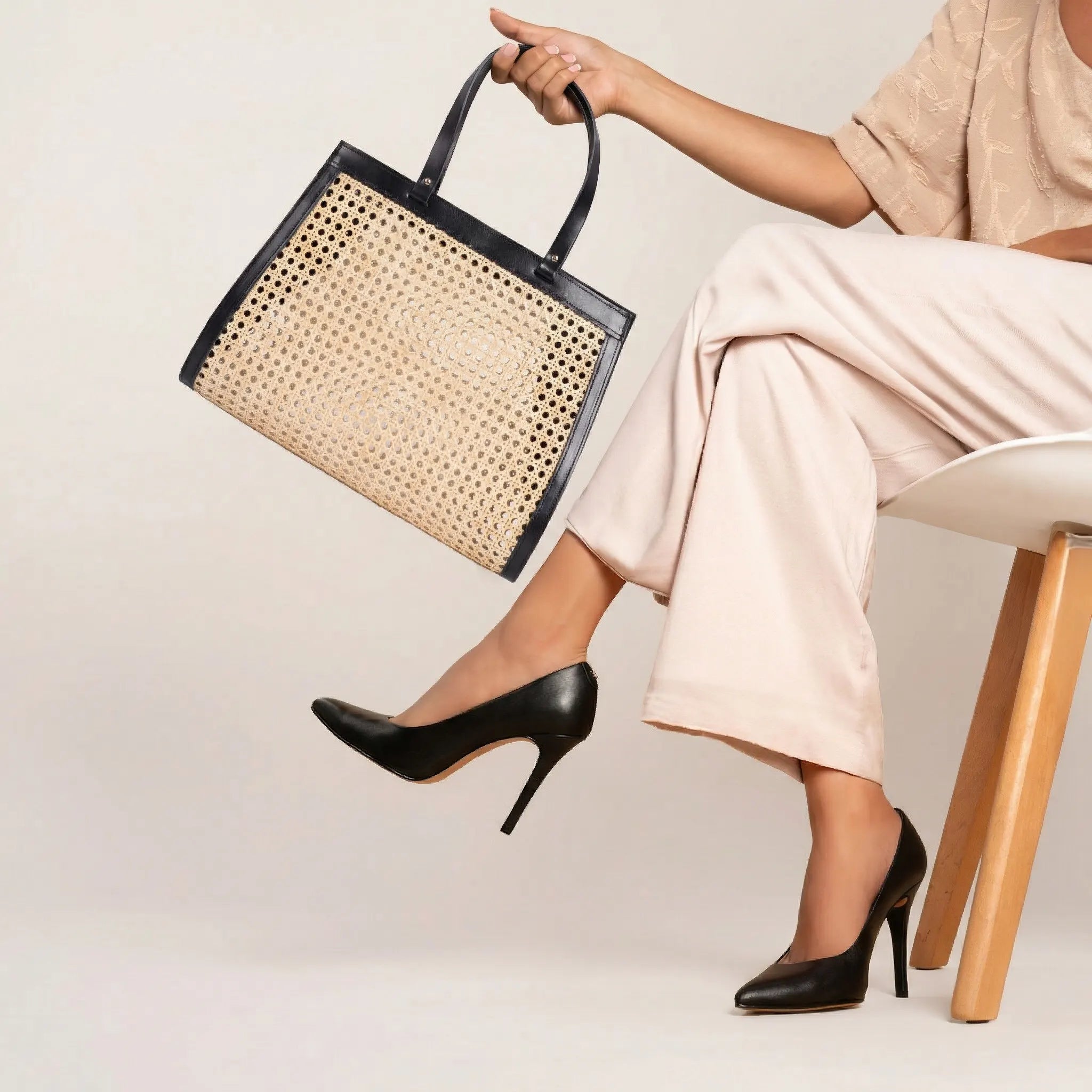Model seated, holding the Lamia rattan cane tote with black leather handles