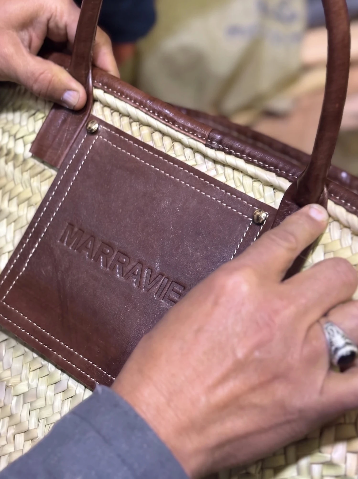 A close-up of a woven palm leaf tote bag with dark brown leather handles and a matching leather front patch embossed with the brand name 'MARRAVIE'. A person's hands are holding the bag in a workshop setting.