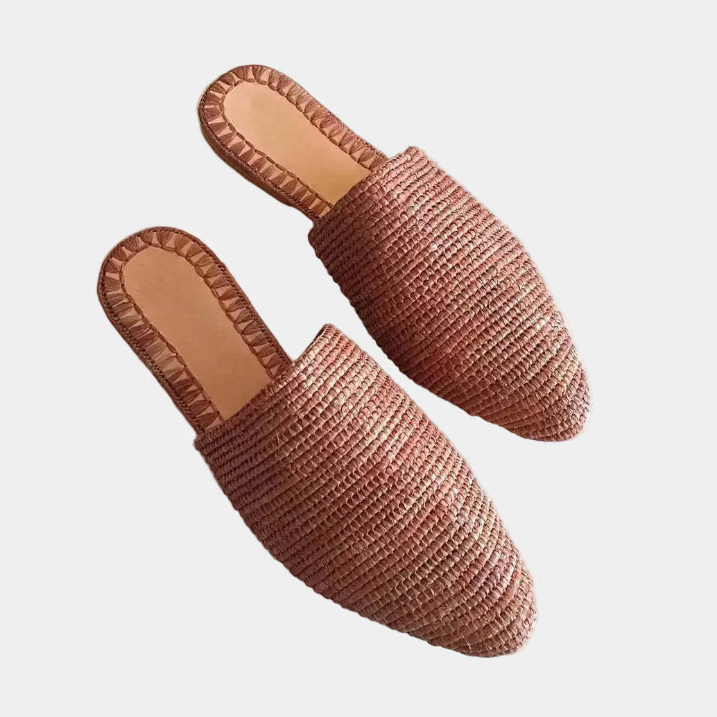 A pair of woven straw mule slippers in a warm terracotta or rust-brown color, shown on a plain white background.