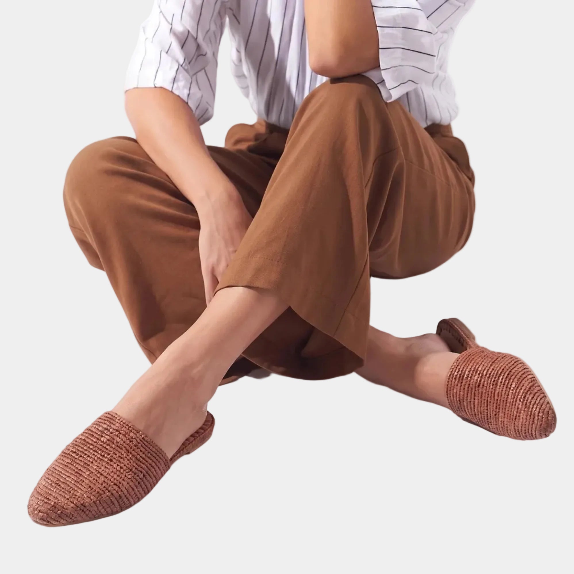 A woman seated on a white floor wearing brown wide-leg cropped pants, a white striped shirt, and textured slip-on shoes in a warm rust-brown color.