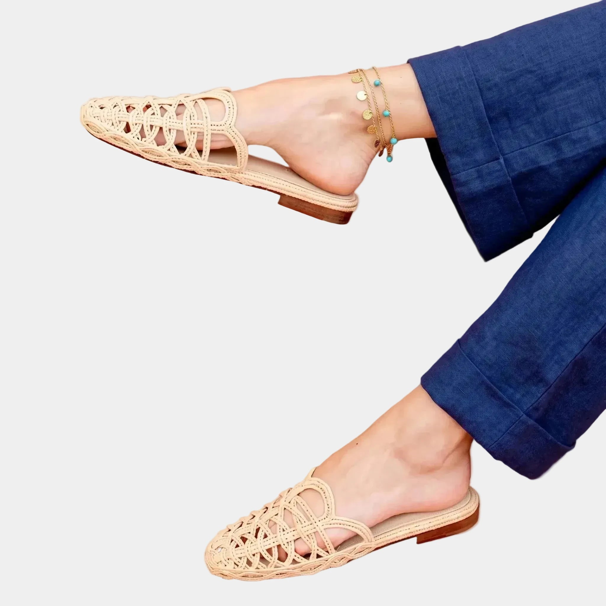 A woman wearing beige woven raffia sandals with blue pants, posed against a solid terracotta background. The sandals feature an intricate open-weave pattern across the footbed and toe area.
